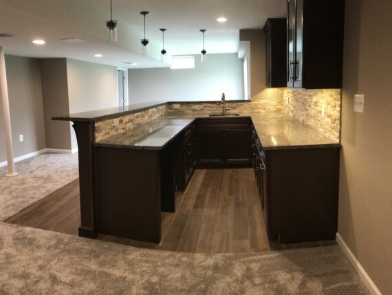 Basement bar with dark cabinetry, stone backsplash, and carpeted flooring.