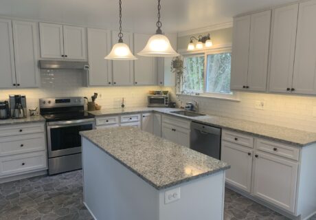 A modern kitchen featuring white cabinets, black countertops, and a stainless steel refrigerator, with pendant lights hanging over a black island.