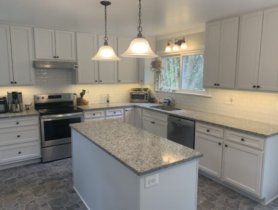 A modern kitchen featuring white cabinets, black countertops, and a stainless steel refrigerator, with pendant lights hanging over a black island.